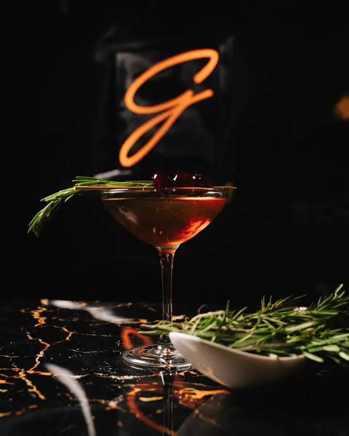 The Autumn in Manhattan, an amber cocktail in a coupe with cherries and a sprig of rosemary, under the neon G.