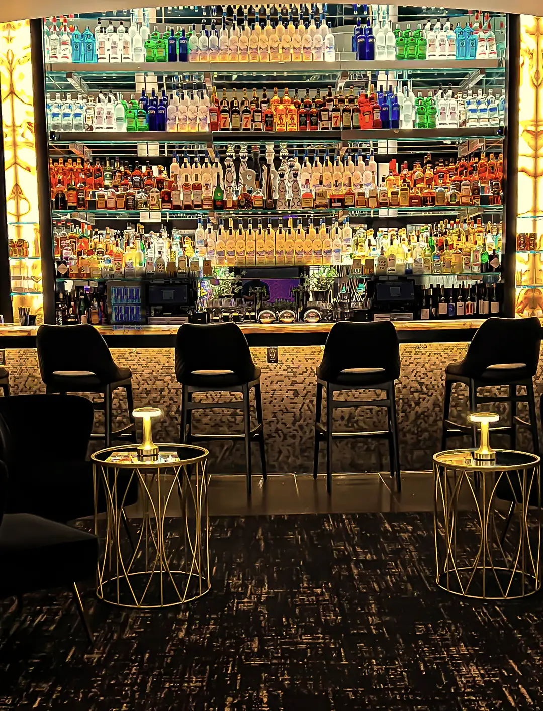 The bar: a backlit wall of bottles between onyx columns, bar stools, and two gold table lamps glowing in the lounge in front.