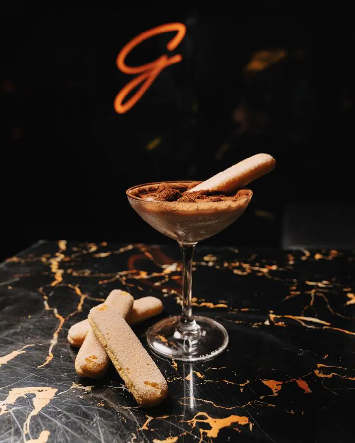 The Tiramisu espresso martini in a coupe, dusted with cocoa, a ladyfinger on the rim and three more on the marble, under the neon G.